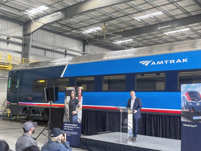 Amtrak President Roger Harris speaks at a podium in front of a new Amtrak Airo train