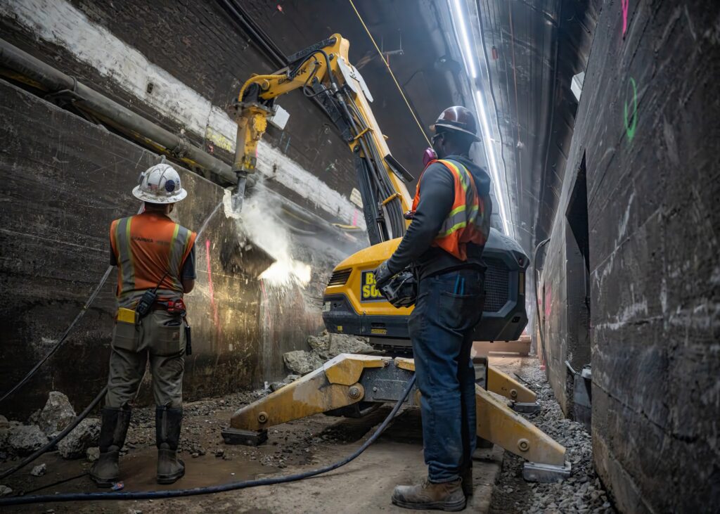 Two workers in a tunnel using construction equipment to demolish the East River Tunnel bench wall