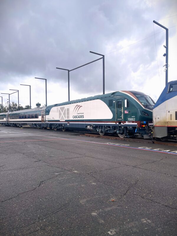 An Amtrak Cascades train sits on outdoor tracks, showing a green‑and‑white locomotive with passenger cars behind it, light poles alongside the platform, and overcast clouds above.