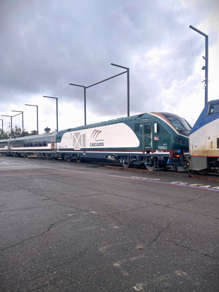 An Amtrak Cascades train sits on outdoor tracks, showing a green‑and‑white locomotive with passenger cars behind it, light poles alongside the platform, and overcast clouds above.