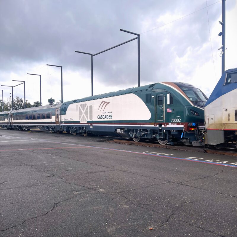 An Amtrak Cascades train sits on outdoor tracks, showing a green‑and‑white locomotive with passenger cars behind it, light poles alongside the platform, and overcast clouds above.