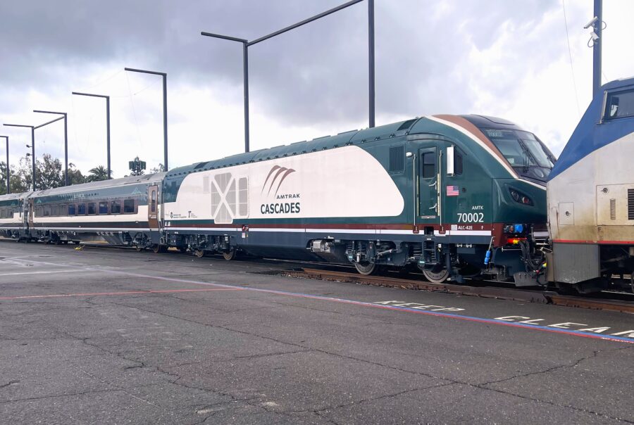 An Amtrak Cascades train sits on outdoor tracks, showing a green‑and‑white locomotive with passenger cars behind it, light poles alongside the platform, and overcast clouds above.