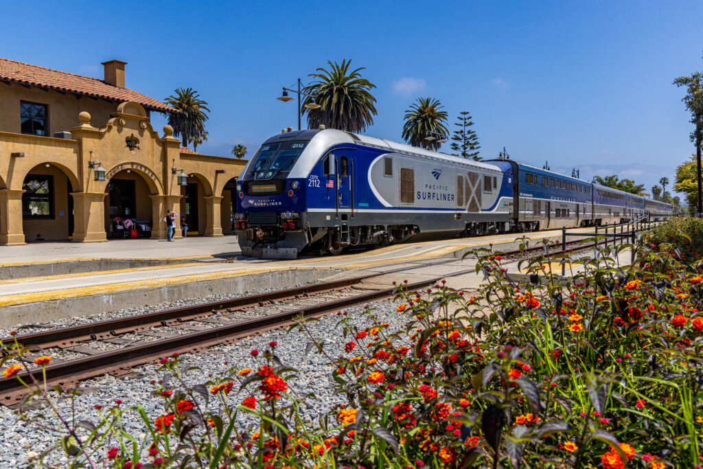 An Amtrak Pacific Surfliner passenger train pulls into Santa Barbara station, framed by palm trees, a historic Spanish-style station building, and vibrant flowers alongside the tracks.