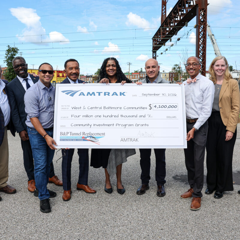 Multiple VIPs representing Amtrak, Congress, and state and local elected officials holding a large check committing 4.1 million dollars to the West and Central Baltimore communities